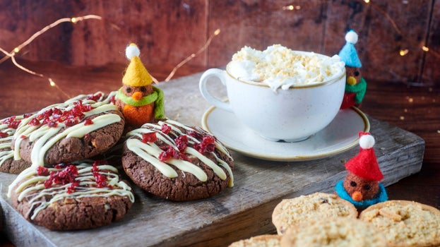 Four chocolate cookies with white chocolate drizzled on top with dried cranberries sprinkled over sit on a board with a festive drink in a cup and saucer. There are some mince pies in the foreground with wire lights behind and three felt birds in bobble hat and scarves sitting amongst it all